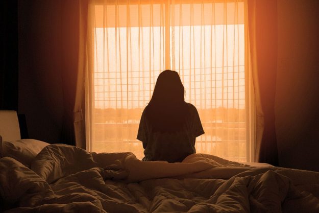 What Causes Bipolar Disorder a silhouette of a person sitting on a bed framed by a large window streaming light while wondering what causes bipolar disorder