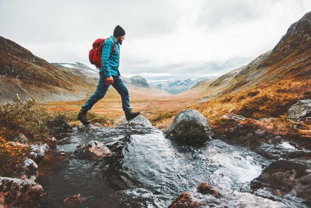 a person in a hat and backpack hikes through a stream in the mountains while wondering what is recreational therapy