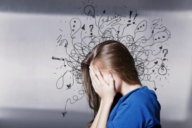 6 Common Causes of Anxiety a person puts their face in their hands in profile with a drawing of squiggles coming out of their head to show the common causes of anxiety