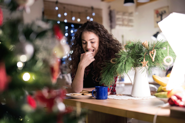 Navigating-Holiday-Triggers–A-Guide-to-Mental-Health-and-Sobriety A woman eats cookies in front of the Christmas tree while she takes a break from her family. She's navigating holiday triggers.